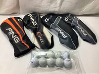 Four Ping golf club head covers and a bag of used golf balls displayed on a white surface.