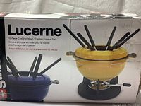 Box of Lucerne 10-piece cast iron meat/cheese fondue set showing yellow and blue color variants on packaging.