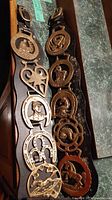 Two leather belts laid out side by side on a dark surface, each with six attached brass horse brasses showcasing detailed equestrian themes.