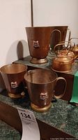 Photo showing three copper cups with handles and three miniature copper kettles on a surface, all exhibiting tarnish and patina.