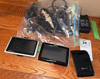 Photo showing Garmin Nuvi GPS, Magellan Roadmate GPS, Blue Ant transponder, and plastic bag with multiple cords, connectors, and chargers on wooden surface.