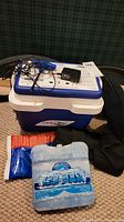 View of blue and white health cooler labeled 'DONJOY' with attached power adapter and cords on top. Two ice packs, one blue and white, one in orange packaging, placed in front of cooler.