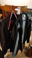 Two black Adidas jackets hanging on a wooden chair, showing the front and back with three white stripes on sleeves.