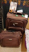 Photo showing brown roller suitcase, matching travel bag, inflatable neck pillow in packaging on top, and a power converter.