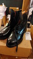 Pair of black men's leather boots size 12 on a cardboard box marked 12, laces undone and standing upright.