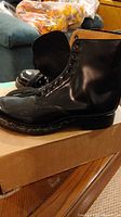 Side view of boots on a box, showing glossy black leather and multiple eyelets for lacing.