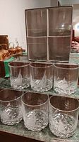 Set of 10 clear drink glasses in original box and displayed on counter showing etched sailboat designs and textured bases.