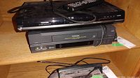 Two black Toshiba electronic devices stacked: DVD player on top and VCR below, with power cables and remote control placed on top, dusty and used condition.