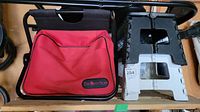 Photo of a black metal folding seat frame with an attached red fabric storage bag labeled 'The Beer Store' next to two plastic folding step stools, one black and one white.