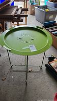 Green metal round side table displayed in a garage with other furniture. Shows the round perforated floral design tabletop with raised edge and handles.