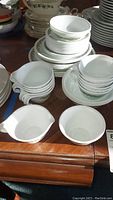 Stack of white Corelle dishes, bowls, and cups with green floral decorative border on wooden surface