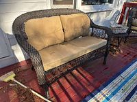 Dark brown woven wicker outdoor couch with beige cushions on a wooden porch floor. Cushions show staining and wear.
