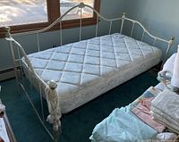Full view of single day bed with mattress on green carpet, cream metal frame with gold ball finials at corner posts.