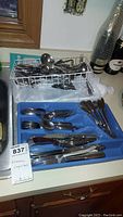 Blue plastic flatware organizer tray with multiple forks, spoons, and knives, and wire basket holders with additional spoons visible.