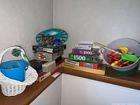 Stack of board games and puzzles on a wooden shelf including Monopoly, Memory game, LIFE, and 1000-1500 piece puzzles. Also includes white basket with blue beach toys.