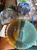 Display of various serving dishes including pottery bowl, Asian themed blue and white covered ceramic dish, glass serving dish, small green leaf dish, two black glass containers, and small white square dish on wooden table.