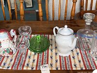 Wide shot on wooden table showing all items including Santa teapot, uranium green glass dish with lid, champagne glasses, white ceramic teapot, clear glass decanter, and tall clear glass.
