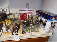 Photo shows boxed Coca Cola hotdog and bottle toaster, ceramic oil and vinegar dispensers with fruit design, sugar jar, glass decanters, various kitchen utensils, and boxed knife set.