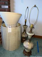 Two matching vintage lamps with metal bases and floral painted glass bodies, with four non-matching lamp shades stacked inside and beside a drum shaped container.