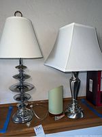 Three lamps shown side by side on a table against a wall: two metal base lamps with white fabric shades and one small lamp with pale green cloth shade.