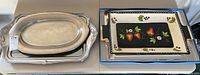 Four serving platters shown together, two metal oval platters and two ceramic rectangular platters with colorful fruit designs.