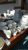 Multiple stacks of white Royal Wentworth china with floral green and gold trim on a wooden table, showing plates, bowls, cups, and sugar bowl with lid.