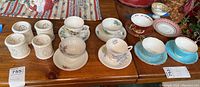 Photo showing 8 assorted bone china tea cups and matching saucers on wooden table including colors white, cream, pastel blue, and floral patterns