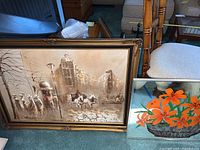 Photo showing the signed painting of a street scene with figures, horse carriage, and buildings in a brown ornate frame, as well as the mirrored wall art with orange flowers in a basket leaning against it.