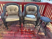 Two dark brown wicker chairs on a wooden deck, each with beige seat and back cushions. One seat cushion is partially torn showing inner stuffing. Small black stool visible nearby.