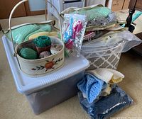 Photo showing wool scarves folded on top of a storage bin with yarn balls in a decorative basket and knitting supplies in plastic bags.