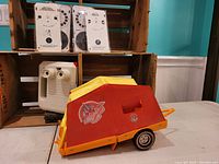 Barbie pop-up camping trailer with red and yellow colors, displayed in front of wooden crates. Visible decals with trailer wheels on table.