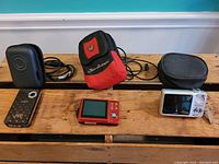 Three digital cameras placed on wooden surface; JVC camera with black case on left, red Centrios camera with red and black pouch in center, and silver Fujifilm camera with black case on right.