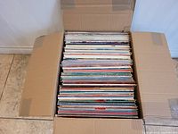 Top-down view of a large cardboard box filled with approximately 100 Christmas vinyl records in sleeves, showing colorful album covers.