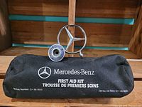Mercedes-Benz first aid kit pouch with black fabric featuring white Mercedes-Benz logo, part number 0 4 86 0033, front view with detached hood ornament behind.