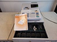 Front view of a Sharp XE-A202A cash register on tabletop with manual placed on top of open cash drawer showing compartments and keys.