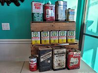 Front view showing all vintage metal cans stacked on a wooden crate, including Coleman Fuel, BP Bar-B-Q Lighter Fluid, Mershol Motor Oil, and CTI Guard Engine Oil cans.