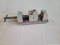Side view of a small vintage metal vice on a white textured surface, showing the screw tightening mechanism and jaws.