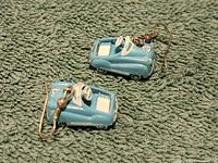 Close-up photo of both blue pedal car-shaped earrings lying on a textured green surface showing detailed design and hooks.