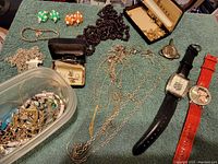 Overview of the jewelry lot showing various chains, watches, vintage earrings, and container with assorted parts on green cloth.