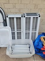 Two white plastic shelves standing upright behind a storage bin containing multiple white plastic support poles. Shelves have ventilation slats and slightly cracked areas.