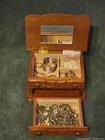 Small open wooden jewelry box showing multiple compartments with various jewelry items inside, including a patterned cuff bracelet, rings, and a plastic bag with small jewelry pieces