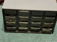Black metal organizer unit with 15 small transparent drawers, some with contents visible, placed on a carpeted surface.