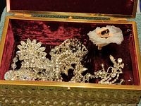 Open jewelry box showing rhinestone jewelry inside against red velvet lining including large floral brooch, bracelet and earrings.
