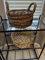 Brown metal 3 tier rack with two decorative woven baskets on shelves in front of window blinds.