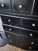 Close-up photo of the black dresser showing three drawers with silver-colored round knobs. Visible scratches and signs of wear on the edges and drawer fronts.