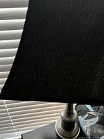 Black table lamp with tapered fabric lampshade and metallic base beside window blinds.