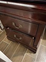 Close-up photo showing the dark cherry wood nightstand with two drawers, both fitted with brass handles. The wood surface looks smooth and polished.