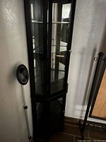 Tall black corner curio cabinet shown placed in corner against wall, featuring glass shelves and glass doors, tall enough to be over 5 feet.