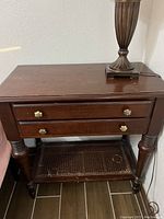 Front angle photo of a wooden Ethan Allen Dawson 2 drawer nightstand with turned legs, lower woven cane shelf and brass round pull handles. Shows surface wear including scratches and water rings.