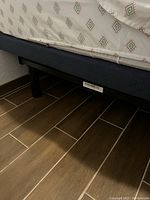 Partial view of black adjustable metal bed frame legs and underside, showing frame height and sturdy construction with mattress edge above on patterned sheet.